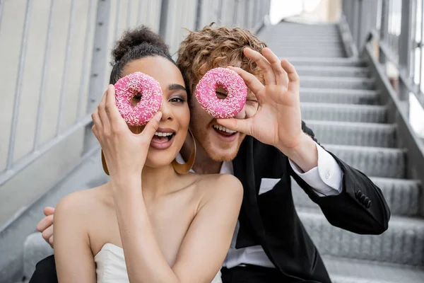 Having fun, wedding, multiethnic newlyweds obscuring face with donuts and looking at camera — Stock Photo