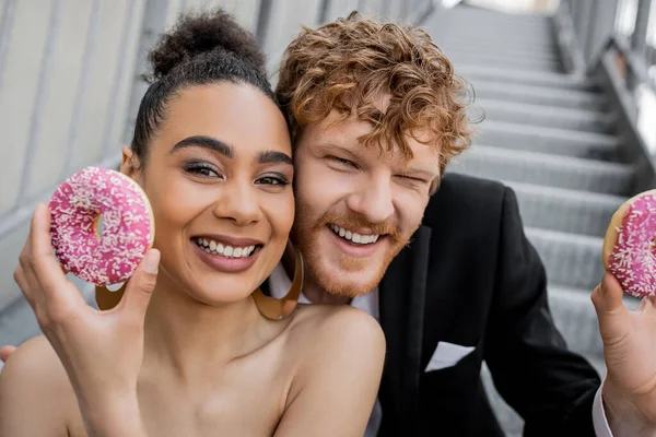 Портрет радостной многонациональной пары с пончиками, смотрящей в камеру, свадьба на городской улице — стоковое фото