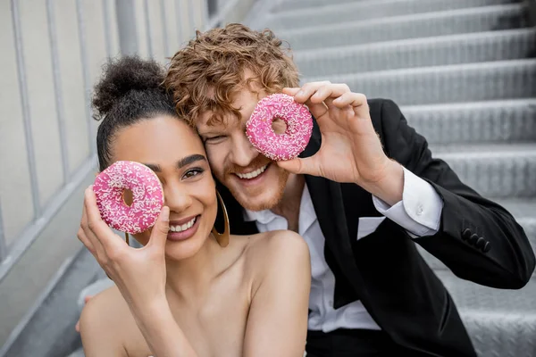 Fun, happiness, wedding outdoors, excited multiethnic couple obscuring face with sweet donuts — Stock Photo