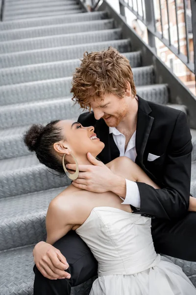 Усміхнений рудий чоловік обіймає молоду афроамериканську наречену на сходах міста, ніжність, щастя — стокове фото