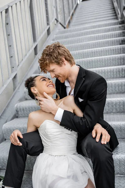 Elegant redhead man embracing delighted african american bride on stairs in city — Stock Photo