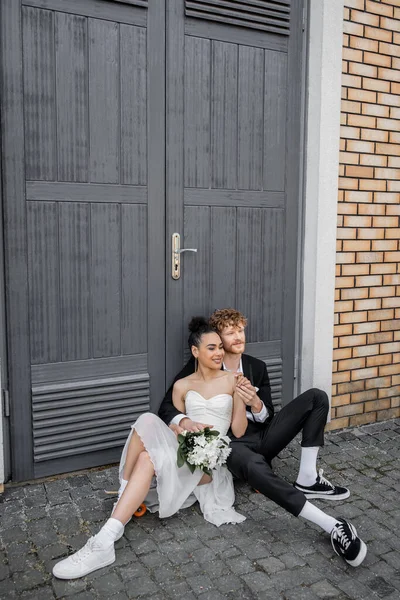 Feliz casal interracial sentado no pavimento da rua perto de portas, traje de casamento, flores — Fotografia de Stock