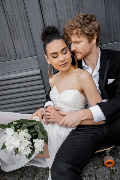 Sposo rossa che abbraccia sposa afro-americana con occhi chiusi e fiori, seduto vicino all'edificio — Foto stock