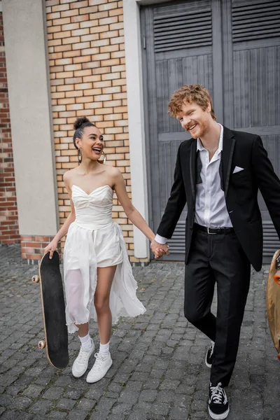 Cheerful interracial newlyweds walking with longboard and skateboard on city street, wedding attire — Stock Photo
