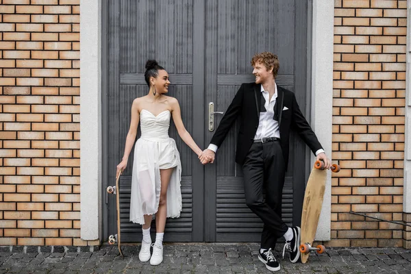 Carefree multiethnic newlyweds with longboard and skateboard holding hands near city building — Stock Photo