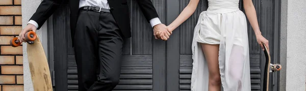 Cropped view of groom with longboard holding hands with african american bride near building, banner — Stock Photo