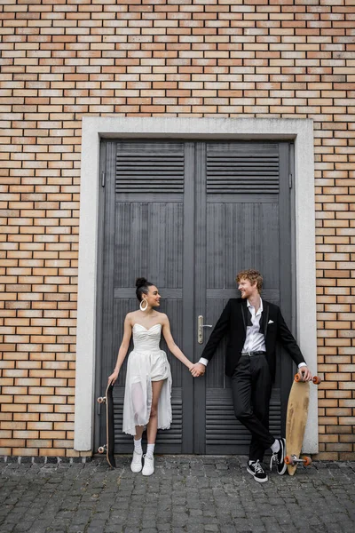 Joyful interracial newlyweds with longboard and skateboard looking at each other near city building — Stock Photo