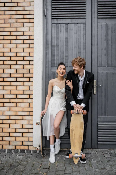Joven pareja interracial con monopatín y longboard cerca de edificio de la ciudad, felicidad, risa - foto de stock