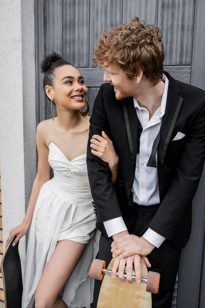 Alegres recién casados multiétnicos con longboard y monopatín sonriendo el uno al otro cerca del edificio de la ciudad - foto de stock