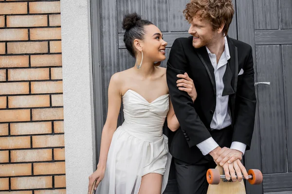 Happy interracial newlyweds with longboard and skateboard smiling at each other near city building — Stock Photo
