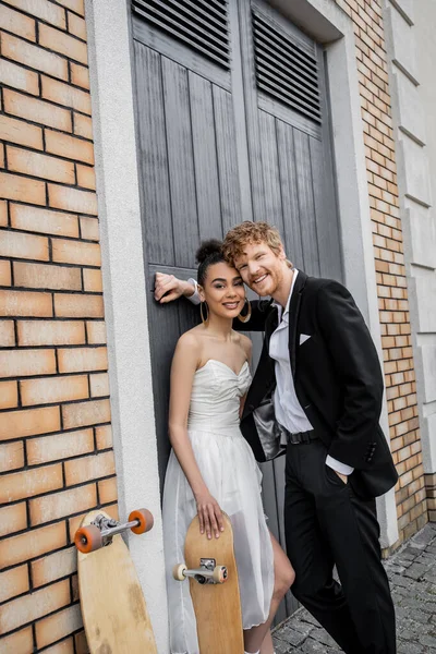 Happy redhead groom with hand in pocket, african american bride with skateboard smiling at camera — Stock Photo