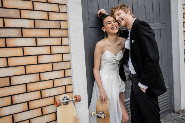 Excited multiethnic newlyweds laughing near longboard and skateboard at urban building — Stock Photo