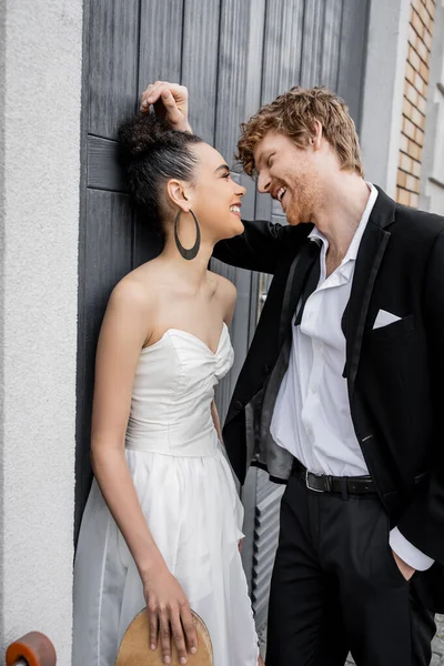 Marié heureux avec la main dans la poche, élégante mariée afro-américaine avec planche à roulettes près du bâtiment — Photo de stock