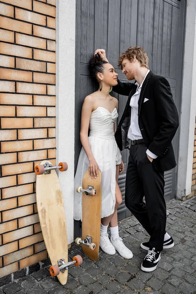 Елегантний рудий наречений з рукою в кишені біля афроамериканської нареченої, довгої дошки і скейтборду — стокове фото