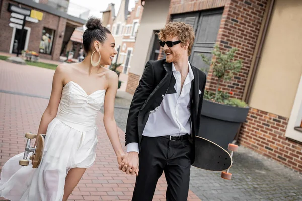 Heureux couple interracial avec longboard et planche à roulettes marche sur la rue, tenue de mariage, lunettes de soleil — Photo de stock