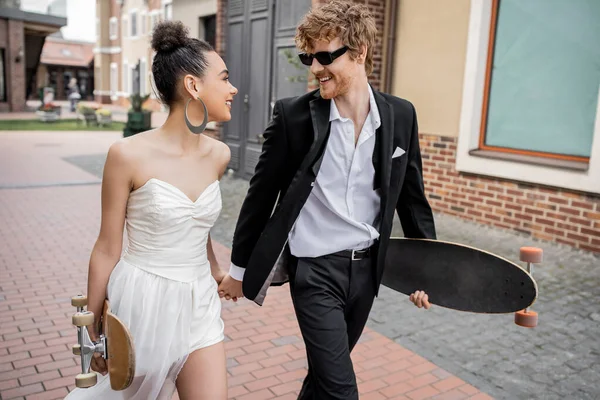 Multiethnic couple walking with longboard and skateboard on street, wedding outfit, sunglasses — Stock Photo