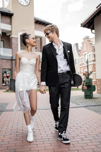 Matrimonio in città, romantica coppia interrazziale a piedi con longboard e skateboard in strada — Foto stock
