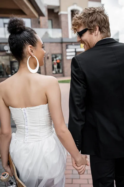 Rossa sposo in occhiali da sole, sposa afro-americana con longboard guardarsi in strada — Foto stock