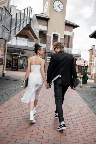 Back view of elegant multiethnic newlyweds walking with longboard and skateboard on urban street — Stock Photo