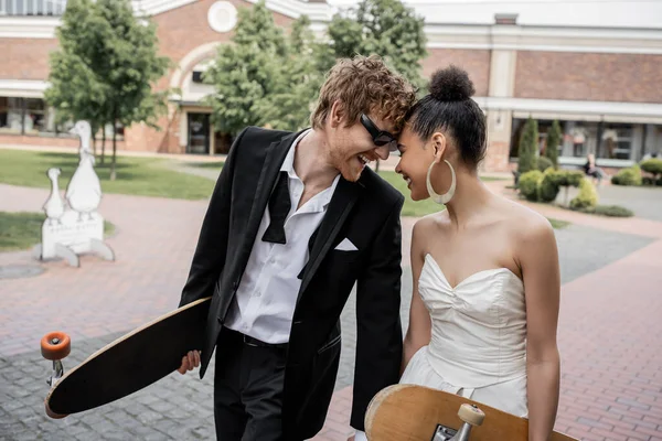 Casal interracial com longboard e skate se divertindo na rua, traje de casamento, óculos de sol — Fotografia de Stock