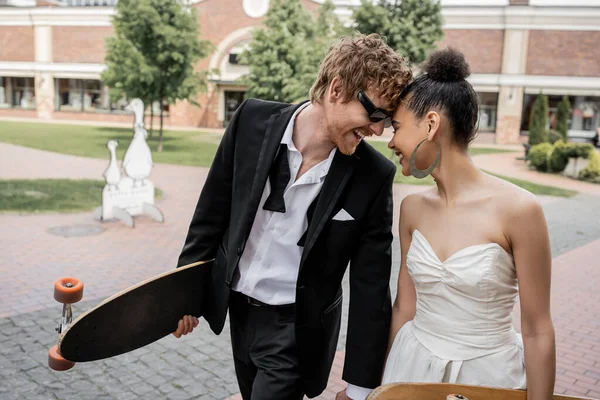 Super alegre multiétnico recém-casados com longboard e skate na rua, óculos de sol, roupa de casamento — Fotografia de Stock