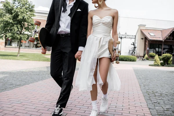 Cropped view of elegant multiethnic couple walking with longboard and skateboard, wedding in city — Stock Photo