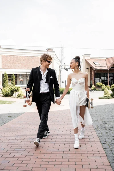 Stylish interracial couple with longboard and skateboard holding, and walking in city, wedding day — Stock Photo