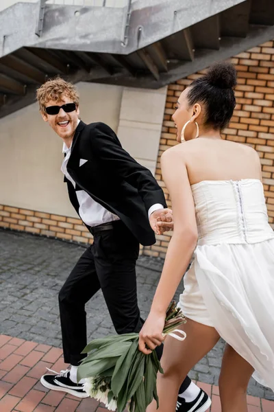 Excitado novio en gafas de sol de la mano de la novia afroamericana y corriendo en la calle urbana - foto de stock