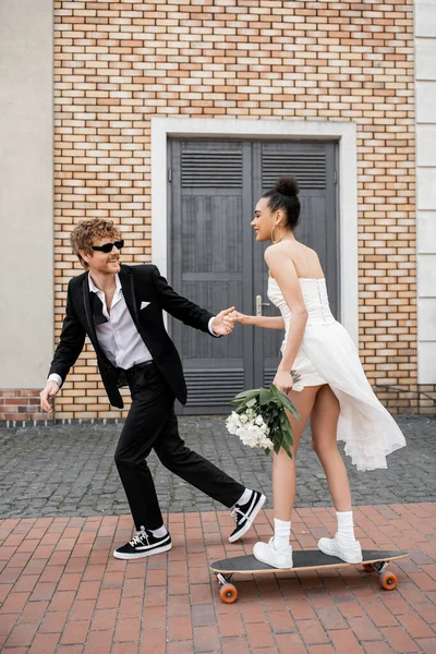 Pelirroja novio en gafas de sol de la mano con la novia afroamericana con ramo de montar longboard - foto de stock