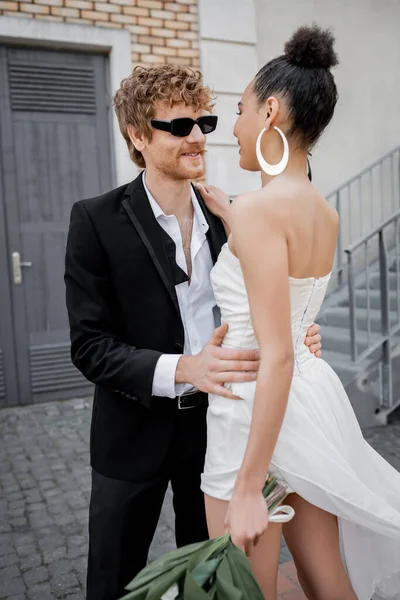 Smiling redhead man in sunglasses embracing elegant african american bride on urban street — Stock Photo