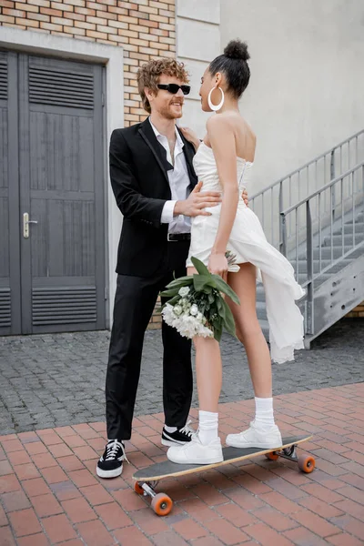 Pelirroja novio en gafas de sol abrazando joven afroamericana novia con flores de pie en longboard - foto de stock