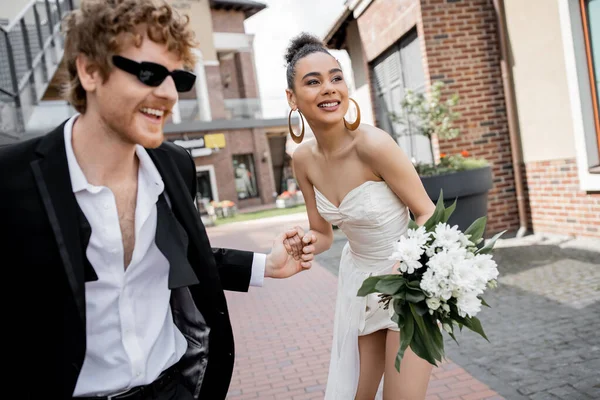 African american american bride with flowers and redhead groom in sunglasses holding hands on street — Stock Photo