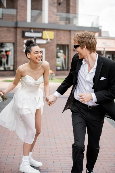 Urban romance, elegant interracial newlyweds holding hands and running in european city — Stock Photo