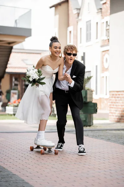 Überglückliche Afroamerikanerin im Hochzeitskleid beim Longboard-Reiten neben elegantem Bräutigam auf der Straße — Stockfoto