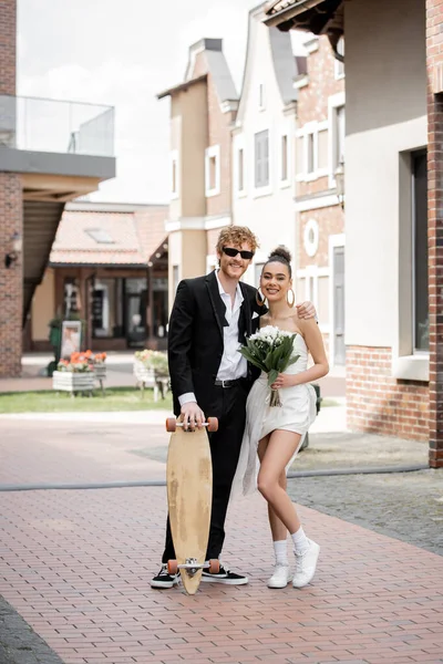 Happy multiethnic couple with longboard and flowers looking at camera, wedding in european city — Stock Photo