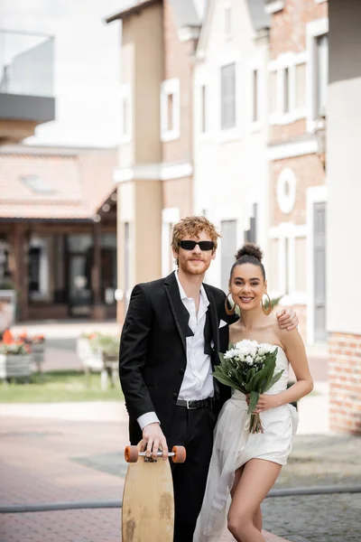 Outdoor wedding, european city, stylish interracial newlyweds with longboard and bouquet on street — Stock Photo
