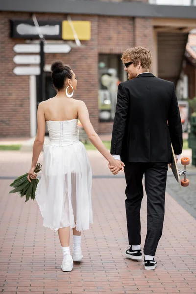Vista posteriore di coppia multietnica con longboard e fiori che si tengono per mano, passeggiando in città, matrimonio — Foto stock