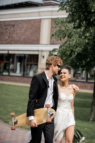 Noivo ruivo com longboard andando com noiva americana africana, traje de casamento, óculos de sol — Fotografia de Stock