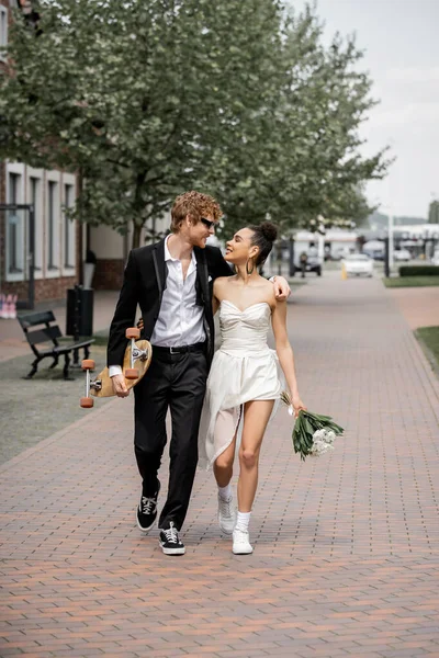Cheerful interracial newlyweds walking with longboard and flowers in european city — Stock Photo