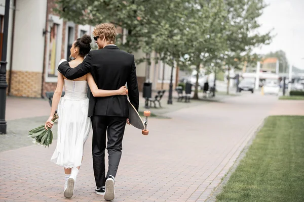 Back view of multiethnic elegant newlyweds with bouquet and longboard embracing and walking in city — Stock Photo