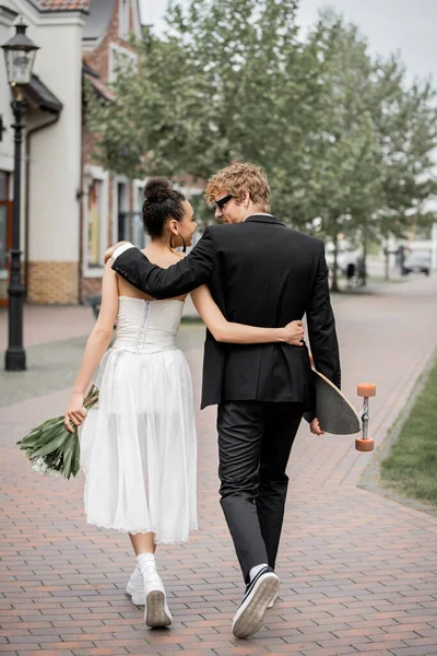 Back view of stylish interracial newlyweds with flowers and longboard embracing and walking in city — Stock Photo