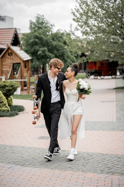 Feliz y elegante pareja multiétnica caminando con longboard y flores, sonriendo el uno al otro - foto de stock