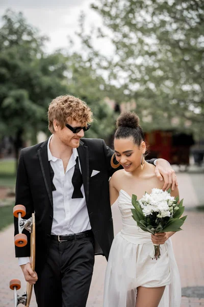 Rothaariger Bräutigam mit Sonnenbrille umarmt afrikanisch-amerikanische Braut, geht mit Longboard und Blumen — Stockfoto