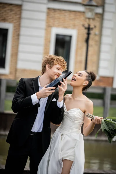 Redhead groom holding champagne bottle near african american bride with bouquet on city street — Stock Photo