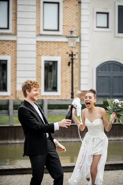 Noiva americana africana animado perto do noivo com champanhe derramando de garrafa, celebração ao ar livre — Fotografia de Stock