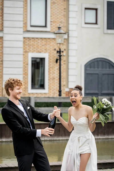 Matrimonio in città, sposo rossa con champagne versando dalla bottiglia vicino urlando sposa afro-americana — Foto stock
