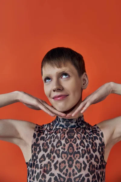 Happy androgynous person smiling and posing on orange backdrop, hands near face, queer fashion — Stock Photo