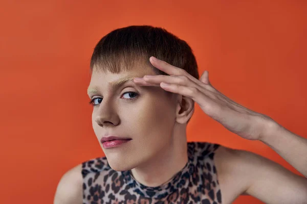 Androgynous person smiling and looking at camera on orange backdrop, touching face, queer fashion — Stock Photo