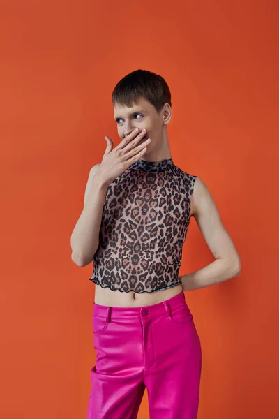 Schüchterne nichtbinäre Person, die lächelt und den Mund auf orangefarbenem Hintergrund bedeckt, queere Mode, rosa Hose — Stockfoto