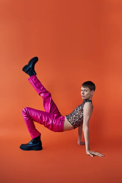 Queer persona en leopardo imprimir sin mangas superior y rosa pantalones posando sobre naranja telón de fondo, moda - foto de stock